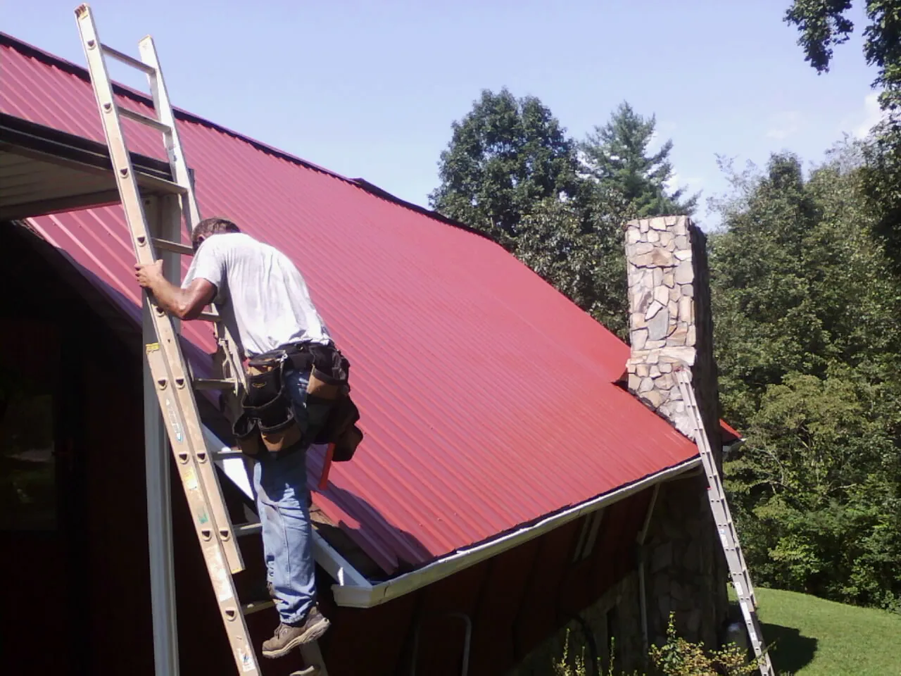Roofer on a ladder beside a red metal roof and stone chimney — Soffit & Fascia Repair by Roofing Brothers in Higden